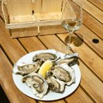 Plate of fresh oysters with a lemon wedge and wine glass on a wooden table outdoors.