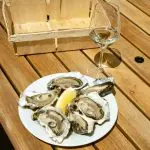 Plate of fresh oysters with a lemon wedge and wine glass on a wooden table outdoors.